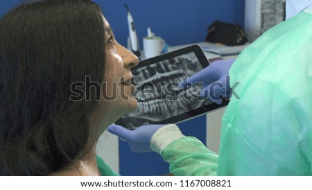 Close up of handsome young dentist showing female patient x-ray on tablet. Dental hygienist and senior brunette woman standing backwards to the camera. Mature caucasian lady turn her face to the