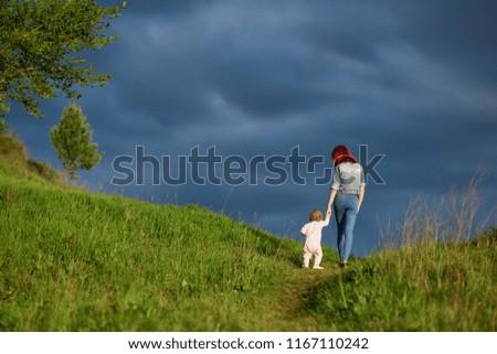 Backview of young mother going with little daughter in summer field on cloudy sky background. Mom and child spending family time together. Woman having red hair, slim figure, wearing jeans clothes.