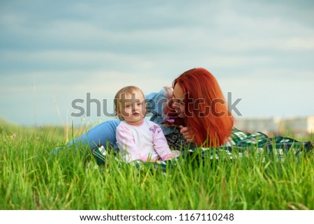Young smiling mother playing with little daughter laying on grass in green summer field, having family time. Woman having red hair, wearing blue jeans, blue shirt. Child looking cute, funny, happy.