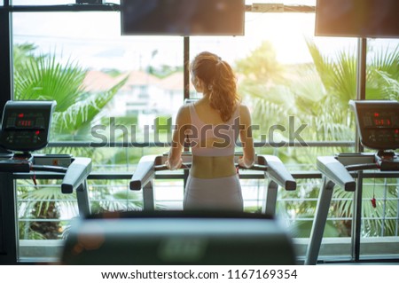 Rear view of fitness woman exercising with listening music and running on treadmill machine in gym in blurred background front of against big window,concept for exercising 