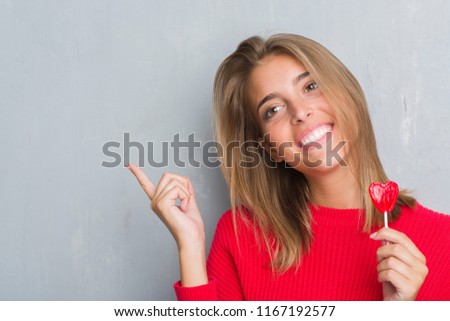 Beautiful young woman over grunge grey wall eating red heart lollipop candy very happy pointing with hand and finger to the side