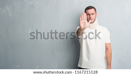 Young caucasian man injured over grey grunge wall wearing neck collar with open hand doing stop sign with serious and confident expression, defense gesture