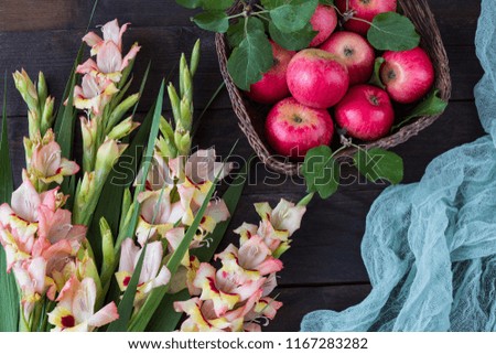 on a dark wooden table in a wicker basket red apples, green gauze and a bouquet of gladioli - summer background