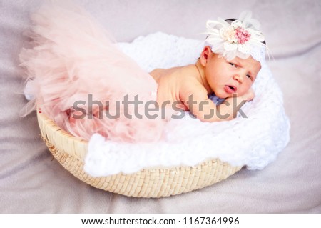 A sweet unhappy to be awaken newborn infant baby girl. A surprised and upset look. A tiny newborn girl in a soft basket studio portrait. Head flower bow on her head. 