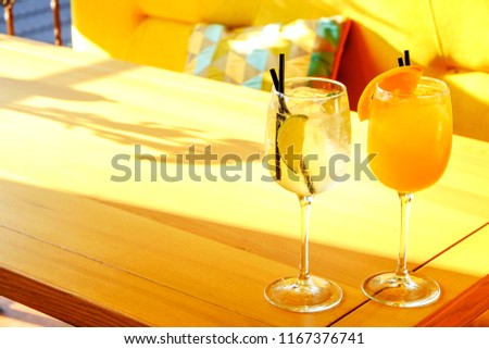 Two glasses of fresh summer non alcoholic refreshing cocktail, ice, orange & lemon on table of patio outside hip restaurant with cushioned seats. Soft sunlight ray. Background, copy space, close up
