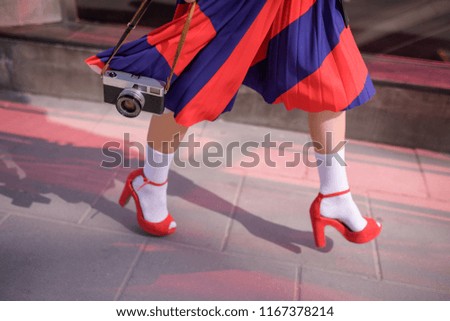 Elegant lady wearing stylish red skirt and shoes holds in hands vintage photo camera. Low angle view of her shapely legs. Selective focus
