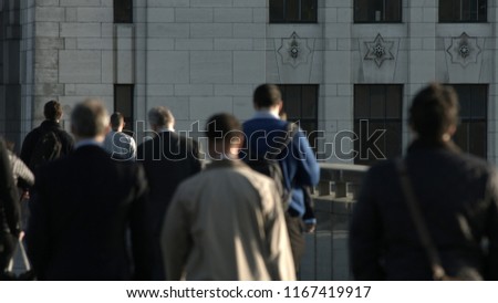 Large crowd of unidentifiable commuters and pedestrians walk across London Bridge to the City of London. In the background is a plain grey building. City workers. Formal clothes. Framed for graphics.