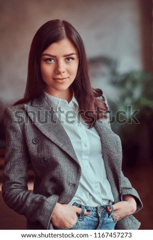 Charming brunette woman dressed in a gray elegant jacket posing with hands in pockets while leaning on a table in a room with loft interior, looking at camera.