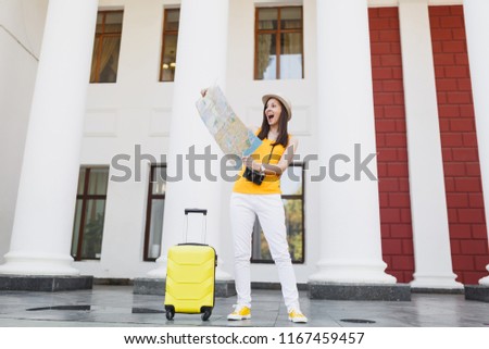 Excited traveler tourist woman in yellow summer casual clothes and hat with suitcase looking on city map in city outdoor. Girl traveling abroad to travel on weekend getaway. Tourism journey lifestyle