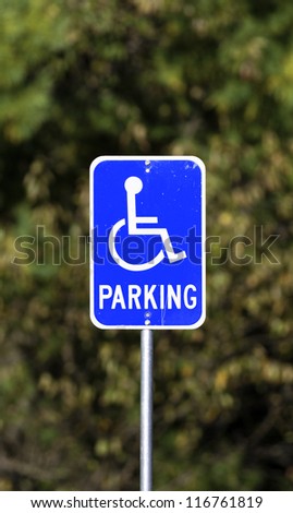 A bright blue handicapped parking sign showing wear with foliage blurred in the background.