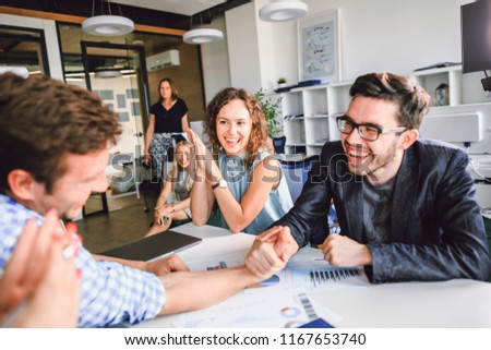 Two men compete arm wrestling at the Desk in the office, the girls support business colleagues clapping their hands