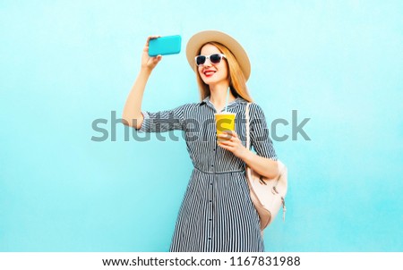 Fashion smiling woman takes a picture self portrait on smartphone holds cup of juice with backpack on blue background