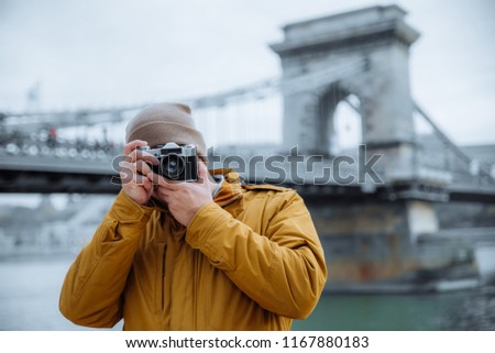 young man in hate taking picture on old retro camera. old chain bridge on background