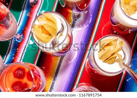 Beautiful layered desserts made of cream, berries and physalis in the tall wineglasses with a spoons standing in a row on a vibrant multicolor tablecloth top angle close-up stylish abstract background