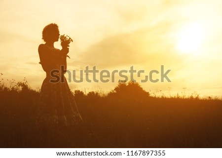 Young woman in dress with bouquet of flowers in hands at sunset in the field. Tinted warm silhouette image