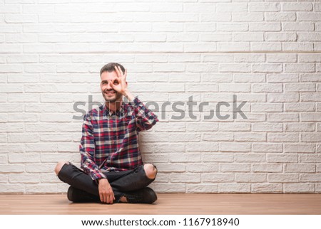 Young adult man sitting over white brick wall with happy face smiling doing ok sign with hand on eye looking through fingers
