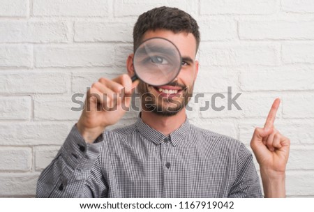 Young adult man over brick wall using magnifying glass very happy pointing with hand and finger to the side