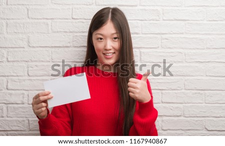 Young Chinese woman over brick wall holding blank paper happy with big smile doing ok sign, thumb up with fingers, excellent sign