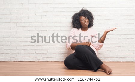 Young african american woman sitting on the floor at home smiling cheerful presenting and pointing with palm of hand looking at the camera.