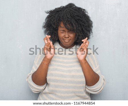 Young african american plus size woman over grey grunge wall wearing a sweater smiling crossing fingers with hope and eyes closed. Luck and superstitious concept.