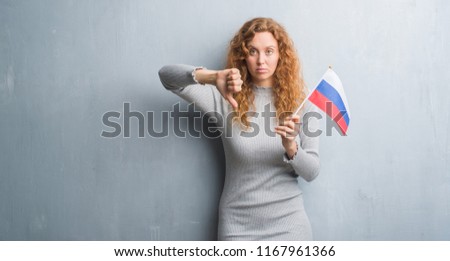 Young redhead woman over grey grunge wall holding flag of Russia with angry face, negative sign showing dislike with thumbs down, rejection concept