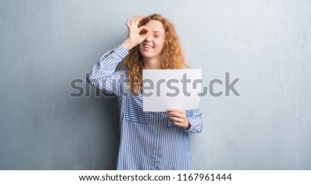 Young redhead woman over grey grunge wall holding blank paper sheet with happy face smiling doing ok sign with hand on eye looking through fingers