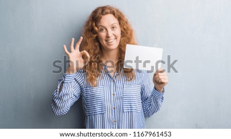 Young redhead woman over grey grunge wall holding blank card doing ok sign with fingers, excellent symbol