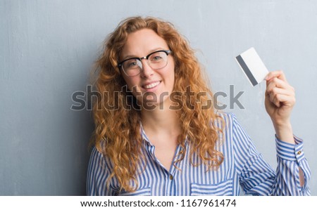 Young redhead woman over grey grunge wall holding credit card with a happy face standing and smiling with a confident smile showing teeth