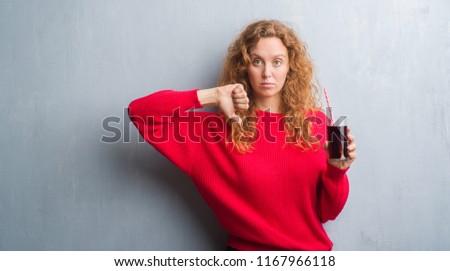 Young redhead woman over grey grunge wall drinking soda refreshment with angry face, negative sign showing dislike with thumbs down, rejection concept