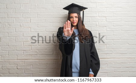 Young brunette woman standing over white brick wall wearing graduate uniform with open hand doing stop sign with serious and confident expression, defense gesture