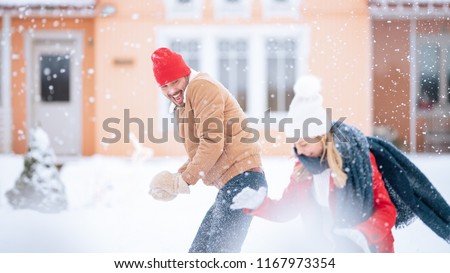 Young Beautiful Couple Throws Snowballs at Each other While Snow Falls. Happy Man and Woman Playing with Snow in the Yard of their Idyllic House. Family Enjoying Winter.