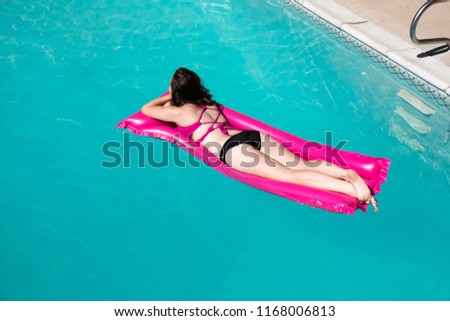 White girl laying on stomach on pink raft. Enjoying lounging in an outdoor swimming pool, blue water and edge of pool. Caucasian girl in bikini on raft in pool.