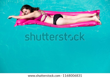 White girl laying on stomach on pink raft. Enjoying lounging in an outdoor swimming pool, blue water and edge of pool. Caucasian girl in bikini on raft in pool arm extended eyes closed.