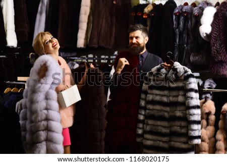 Couple in love: girl in pink fur coat look at striped fur in male hands. Winter clothing and glamour concept. Woman with smiling face in fashion store. Shop assistant with beard shows fur coat to lady