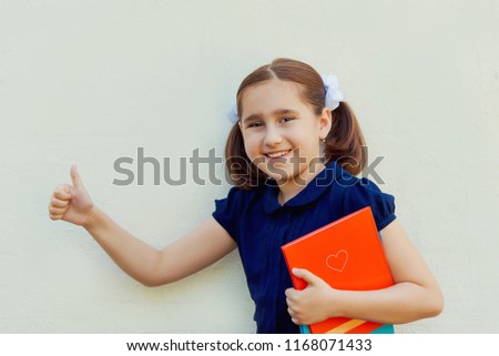 A teenage student with white bows on her head and a book in her hands shows her thumb and smiles against a white background. The concept of a fun educational process.