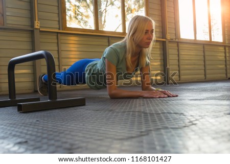 The blonde woman trains on a rubber coating on the background of wall and windows, in the sun, making a bar, fixing the position of the body, leaning his hands on the floor and looking straight ahead