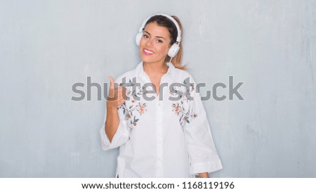 Young adult woman over grey grunge wall listening to music and dancing happy with big smile doing ok sign, thumb up with fingers, excellent sign