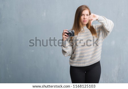 Young adult woman over grey grunge wall taking pictures using vintage camera with angry face, negative sign showing dislike with thumbs down, rejection concept