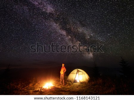 Night camping in mountains. Woman resting near illuminated tent and brightly burning bonfire on deep dark sky with lot of bright sparkling stars and Milky way. Tourism and outdoor activity concept.