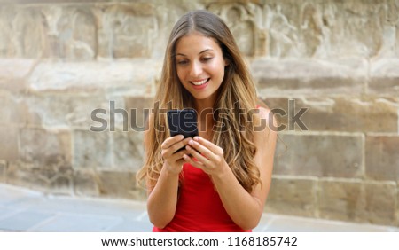 Smiling woman using mobile phone app to play video games online. City woman relaxing. Urban lifestyle. Banner crop for advertising copy space, background european old town stone wall. 