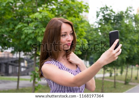 Portrait of attractive young female taking selfie on mobile phone in park. Gorgeous woman posing for picture in striped dress with trees behind. Blurred background