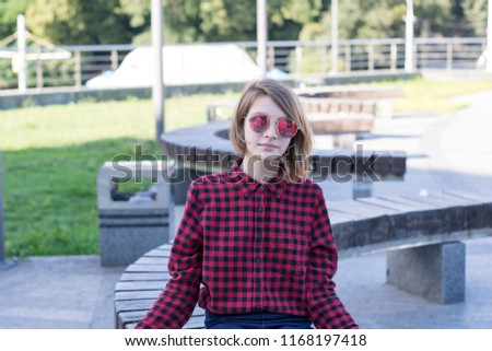 Waist-up portrait of good-looking young female sitting in nature. Well-dressed girl in red plaid shirt and trendy sunglasses. Youth and weekend concept. Blurred background