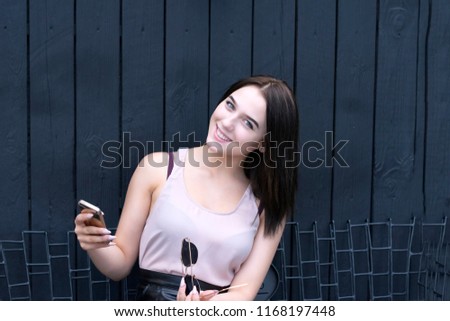 Portrait of smiling young female holding telephone and posing for camera with cheerful mood. Outside picture of cute girl on wooden wall. Technology concept