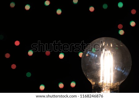 Bare Lightbulb In Foreground And Black Background With Small Lights Showing Bokeh.