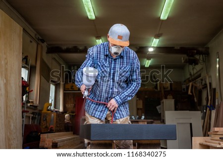 Experienced carpenter in work clothes and small buiness owner   is carving a wooden board on an  modern  hand drill in a light workshop side view, in the background a lot of tools
