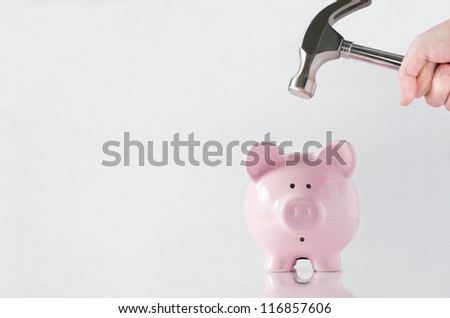 A hand holding a hammer which is raised above a pink china piggy bank, with a shocked and apprehensive facial expression.  Reflective surface and light grey background. Copy space to the left. Royalty-Free Stock Photo #116857606