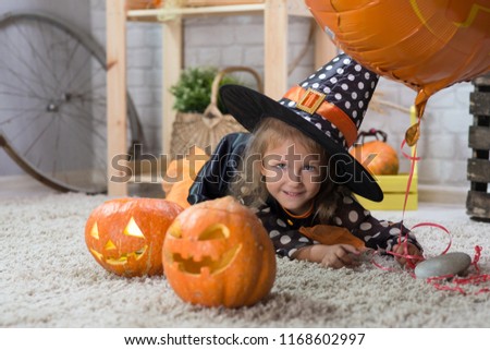 Happy Halloween. A little beautiful girl in a witch costume celebrates a home in an interior with pumpkins