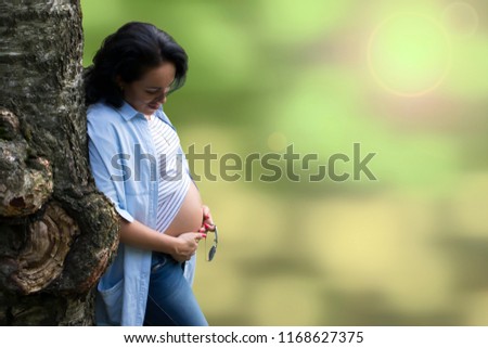 Young beautiful long haired brunette leaning on a bizarre tree and looking at her pregnant belly against blurred bokeh background