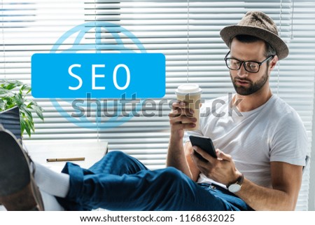 bearded developer in hat holding coffee to go and using smartphone with SEO sign