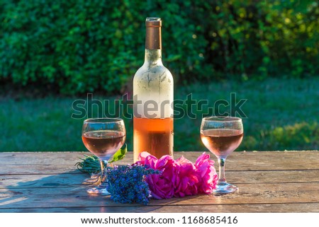 One bottle and two glasses of pink wine, bouquet of roses and lavenders flowers on sunlight on wooden table in the garden